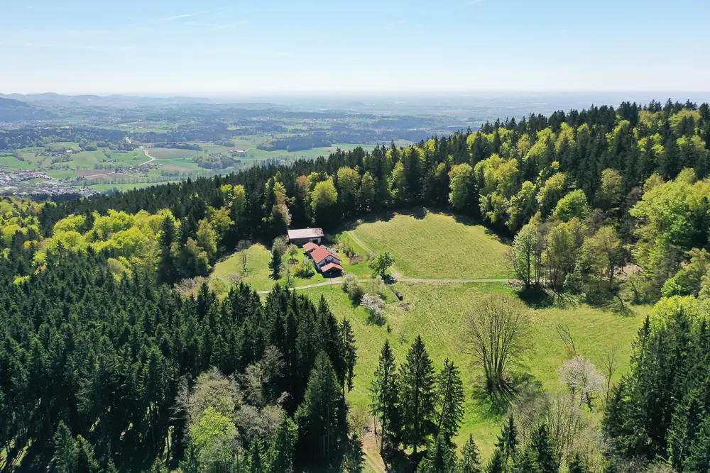 Luftaufnahme: das Bauernhaus Eckhütt Eins als kleiner Punkt auf einer Waldlichtung, umgeben von Bayerischem Wald, mit weitem Blick auf das Tal und die Hügelketten bis zum Horizont.